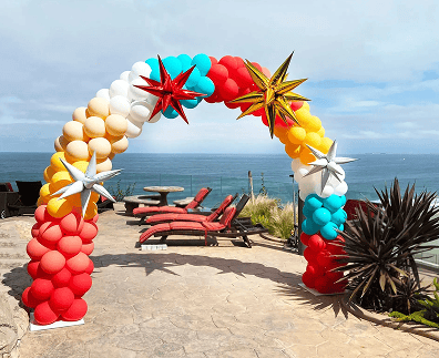 A stunning balloon decor arch in red, yellow, white, and blue with metallic star accents stands on a patio overlooking the ocean, perfect for a corporate event with lounge chairs nearby.