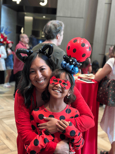 A woman and a girl in matching red outfits with black polka dots pose together indoors in Los Angeles; the girl wears a mask and a balloon hat featuring a ladybug design, adding playful balloon decor to the scene.
