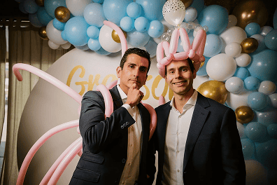 Two men in suits pose in front of a balloon display at a corporate event in Los Angeles; one wears pink balloon wings, the other a pink balloon crown, both standing and looking at the camera—creations by a talented balloon artist.