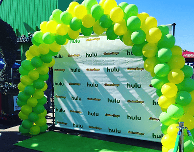 A green and yellow balloon arch stands in front of a backdrop with repeated Hulu and #TheHandmaidsTale logos, showcasing vibrant balloon decor for a corporate event promotional setup.