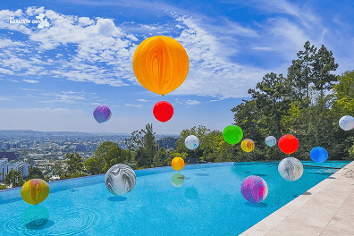 Colorful balloon decor of various sizes floats above an outdoor infinity pool in Los Angeles, offering city views and trees in the background under a partly cloudy sky.