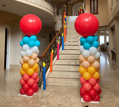 Two large balloon columns, crafted by a skilled balloon artist and each topped with a red balloon, stand on either side of a staircase decorated with colorful candle balloon installations.