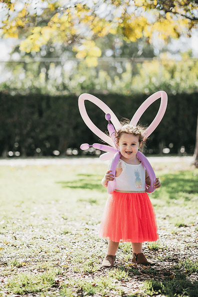 A young child wearing a red tutu and a white top stands outdoors, smiling, with balloon artist-crafted butterfly wing balloons on her back.