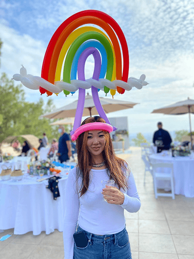 A woman stands outdoors at an event, wearing a colorful balloon hat shaped like a rainbow—crafted by a talented balloon artist—and holding a drink. Tables with food and people are visible in the background.