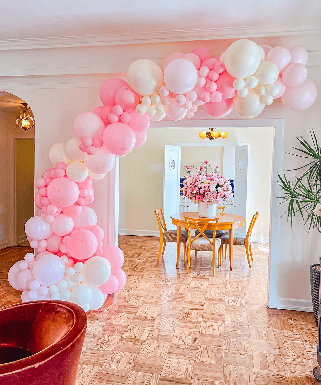 A pink and white balloon arch by a skilled balloon artist decorates a doorway in Los Angeles, leading to a dining area with a round table, wooden chairs, and a large floral centerpiece.