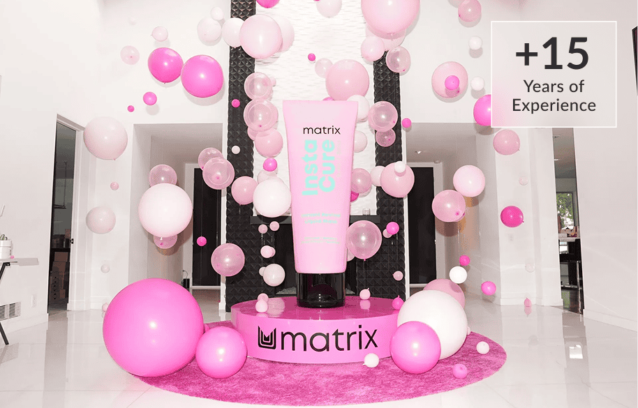Large pink Matrix hair product prop surrounded by pink and white balloons, crafted by a talented balloon artist in Los Angeles, in a bright room; sign reads "+15 Years of Experience." Perfect for a stylish corporate event backdrop.