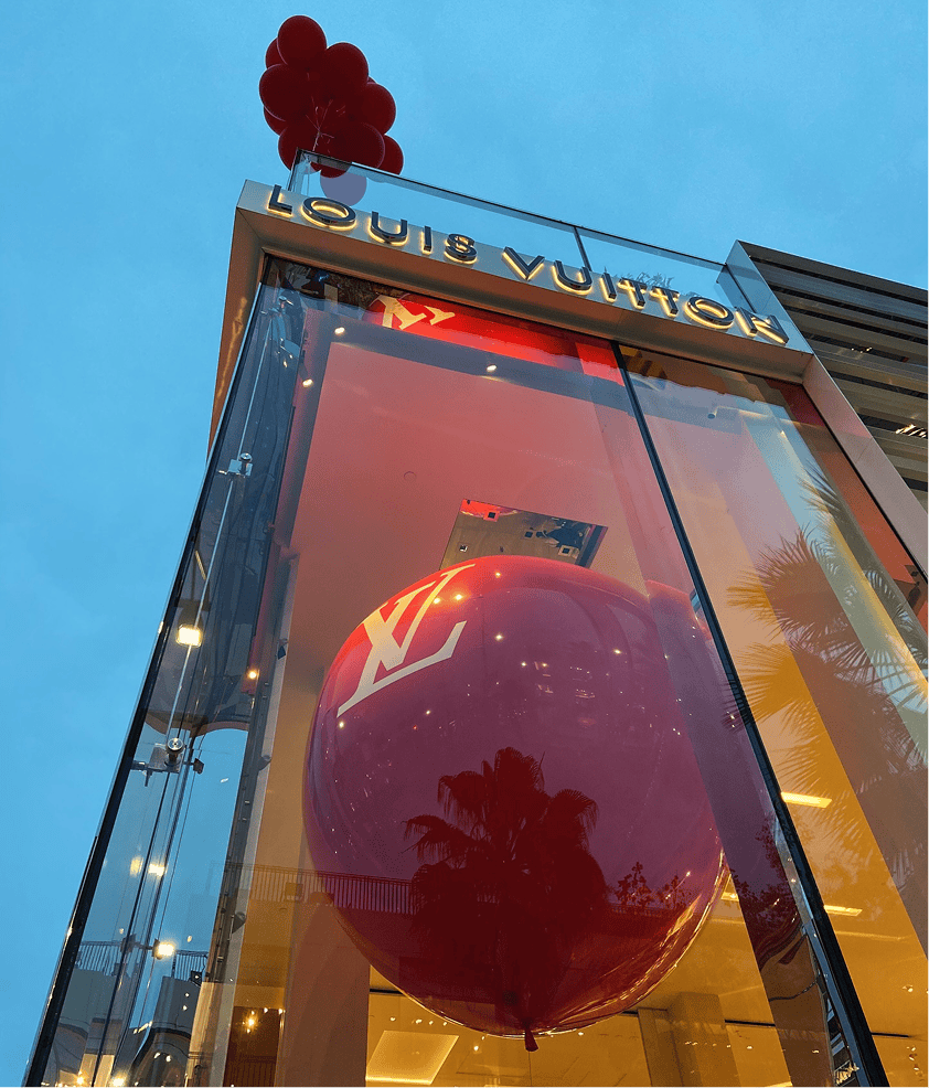 A large red Louis Vuitton balloon installation is displayed inside a glass storefront with the LV logo, while a cluster of red balloons floats above the entrance, creating an eye-catching display perfect for a corporate event.
