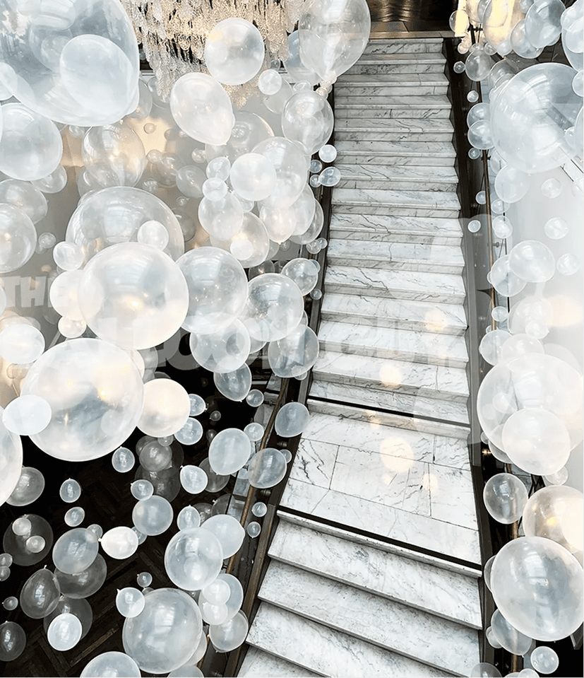 A marble staircase is surrounded by numerous translucent white balloons suspended in the air, creating a whimsical atmosphere reminiscent of elegant balloon installations in Los Angeles.