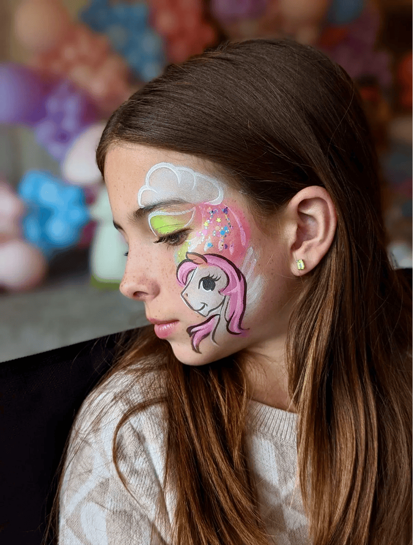Young girl with long brown hair and closed eyes has colorful face paint featuring a unicorn, clouds, and rainbows on her left cheek. She is wearing a light sweater and small earrings, perfect for a day with creative balloon artist fun.