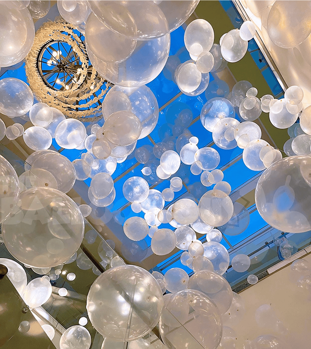 Clear and white balloon-like decorations, crafted by a talented balloon artist, hang from a ceiling with a chandelier and a blue skylight above, creating a floating effect perfect for a stylish corporate event in Los Angeles.