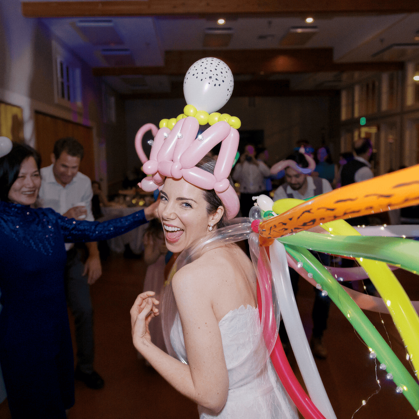 A woman in a white dress smiles while wearing a whimsical balloon hat and colorful balloon wings, showcasing playful balloon decor at an indoor Los Angeles party. Other guests are visible in the background.