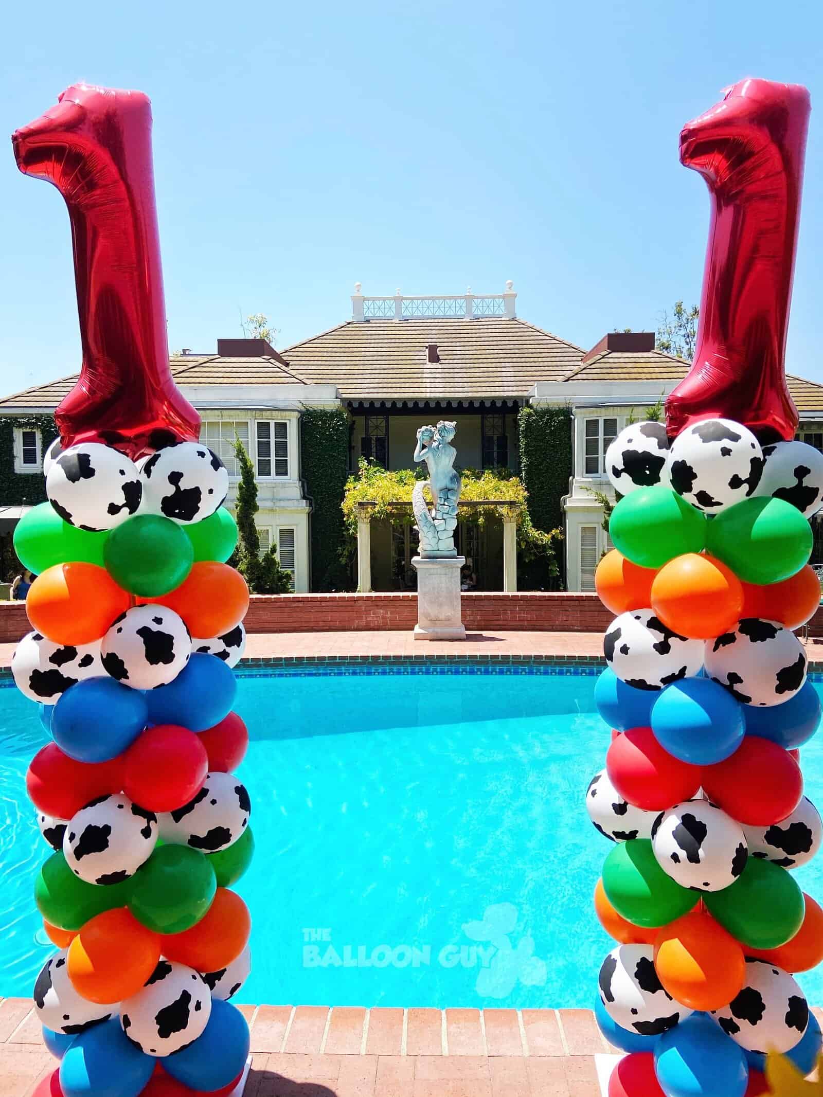 Two balloon columns featuring cow print, orange, blue, green, and red balloons are topped with large red number 1 balloons. Perfect for a corporate event in Los Angeles, they stand by the pool in front of a house.