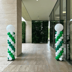 Two Balloon Columns featuring white and green balloons stand beside a walkway by a glass wall and hedge, making eye-catching balloon décor ideal for corporate events.