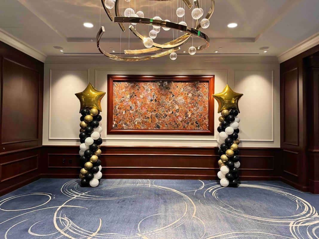 A decorated room features elegant balloon decor by a balloon artist, with two columns of gold, black, and white balloons topped with gold stars, a modern chandelier, abstract artwork, and a blue carpet with circular patterns.