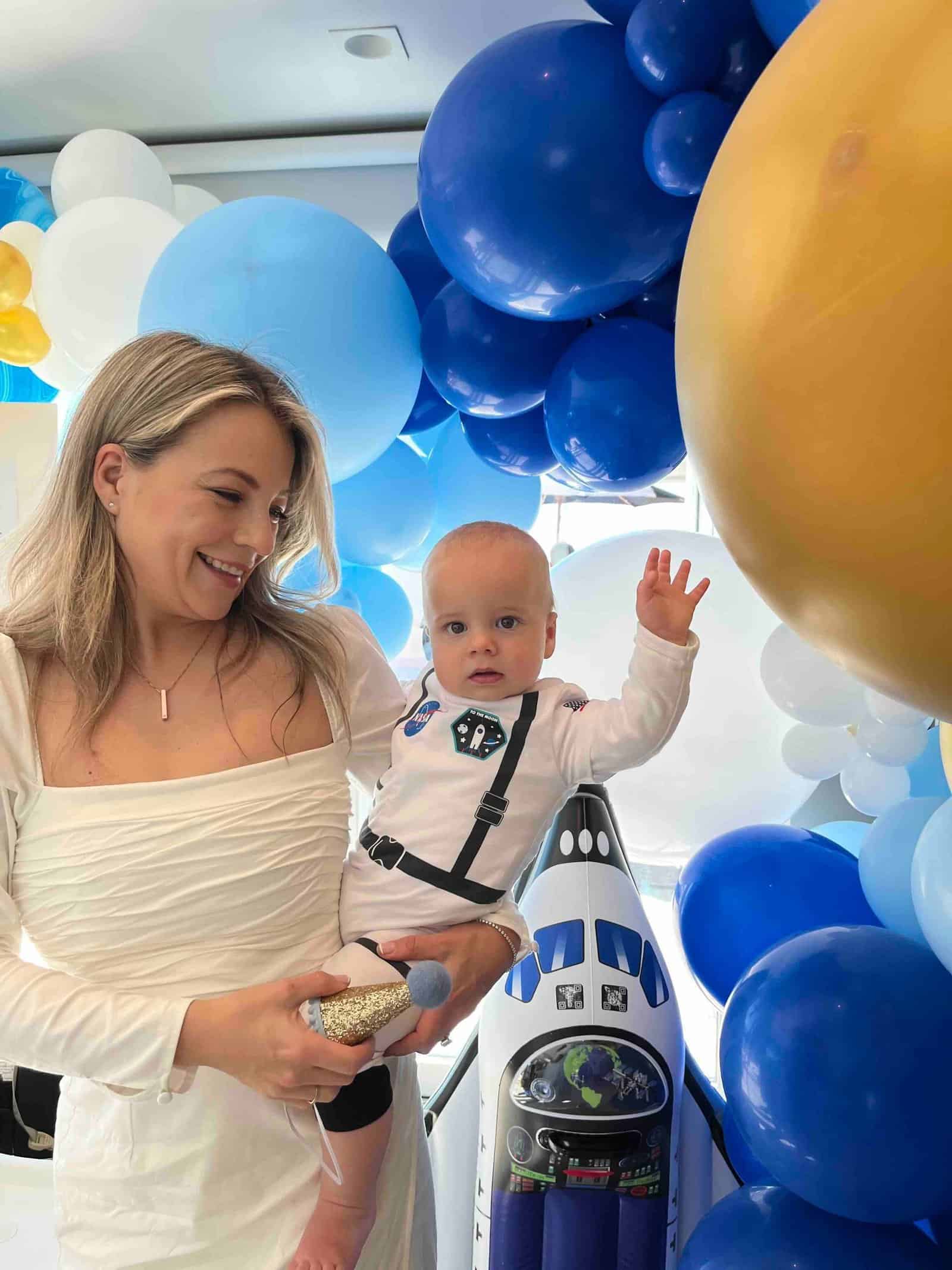 A woman holds a baby dressed in a space-themed outfit, surrounded by stunning balloon installations in blue, white, and gold, with a space shuttle decoration adding to the festive atmosphere.