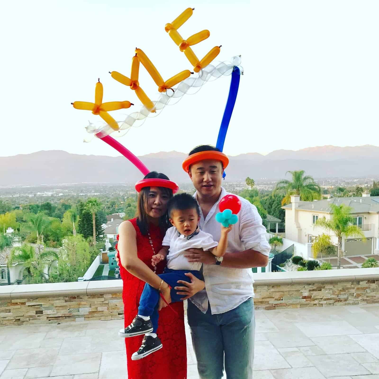A family of three stands outdoors; the parents wear balloon hats crafted by a balloon artist, and the father holds their child. Mountains and houses are visible in the background. Balloon letters spelling "YLE" float above them.