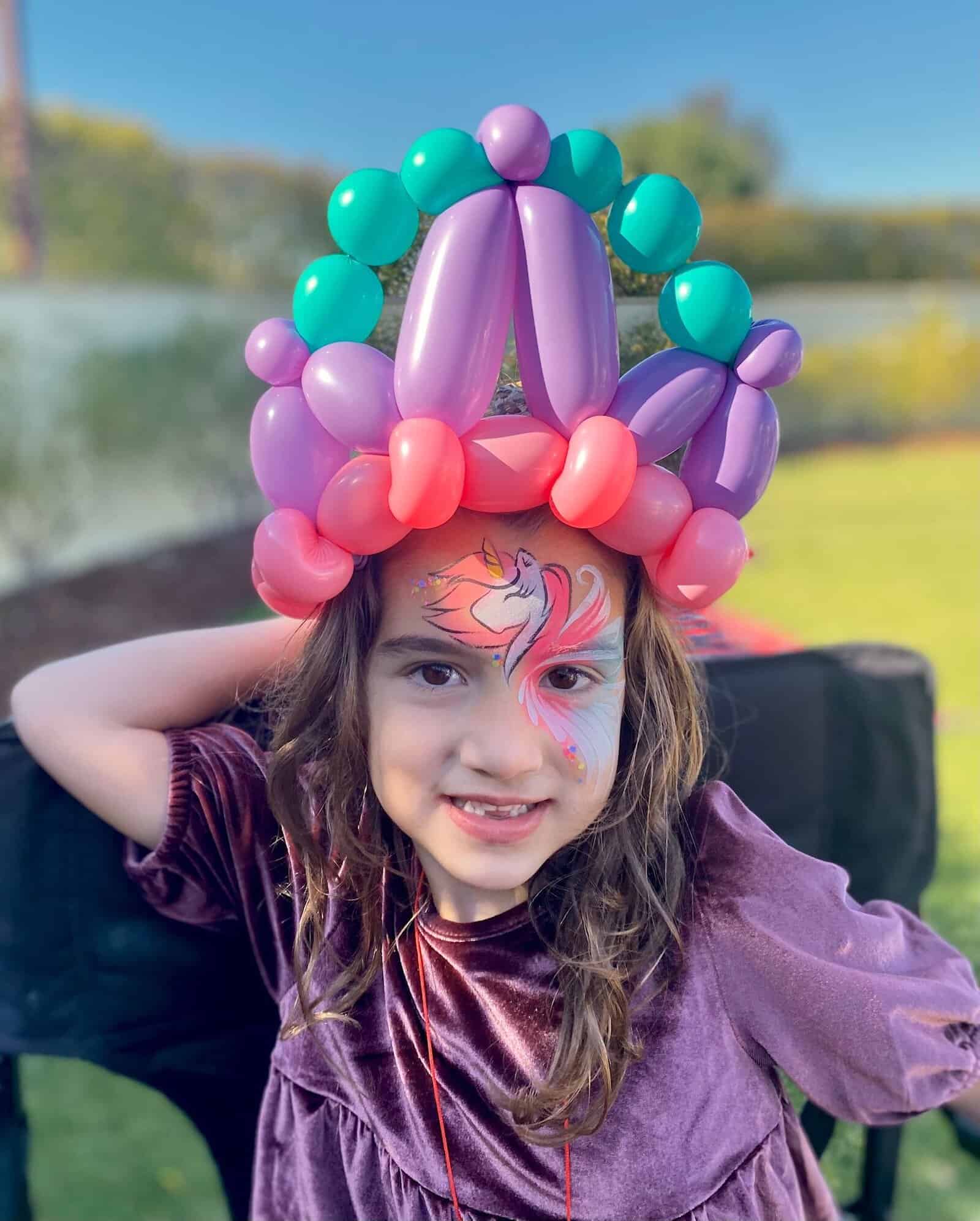A young child wearing a large, colorful balloon crown by a talented Los Angeles balloon artist and a unicorn face painting smiles outdoors, dressed in a purple top.