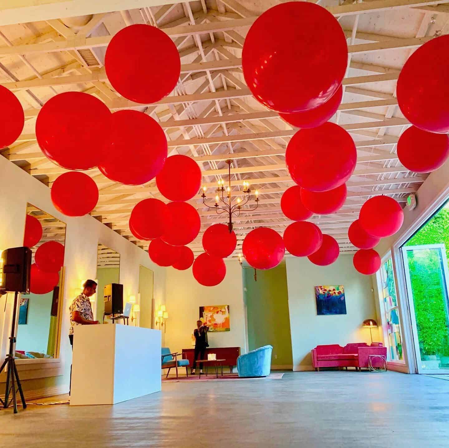 Spacious modern room with high ceiling, striking balloon installations suspended overhead, open glass door, lounge seating, and two people standing by a white counter—perfect for a stylish corporate event.