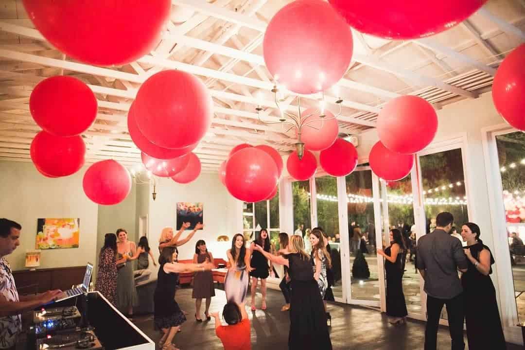 People dance under large red balloons in a well-lit Los Angeles venue with big windows, while a DJ plays music in the corner.