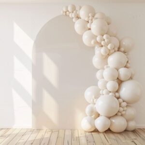 A Chiara Balloon Arch featuring cream-colored balloons of various sizes decorates a white arched wall in Los Angeles, with sunlight creating shadows on the wooden floor.