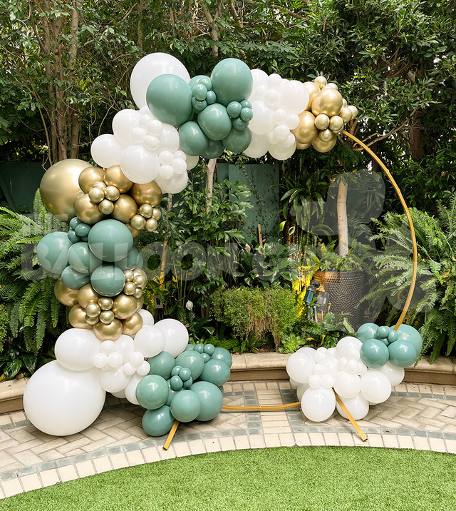 A gold circular frame is decorated with clusters of white, gold, and green balloon decor, set outdoors in front of lush green plants.