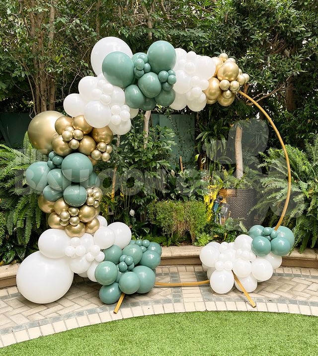 A gold circular frame is decorated with clusters of white, gold, and green balloon decor, set outdoors in front of lush green plants.
