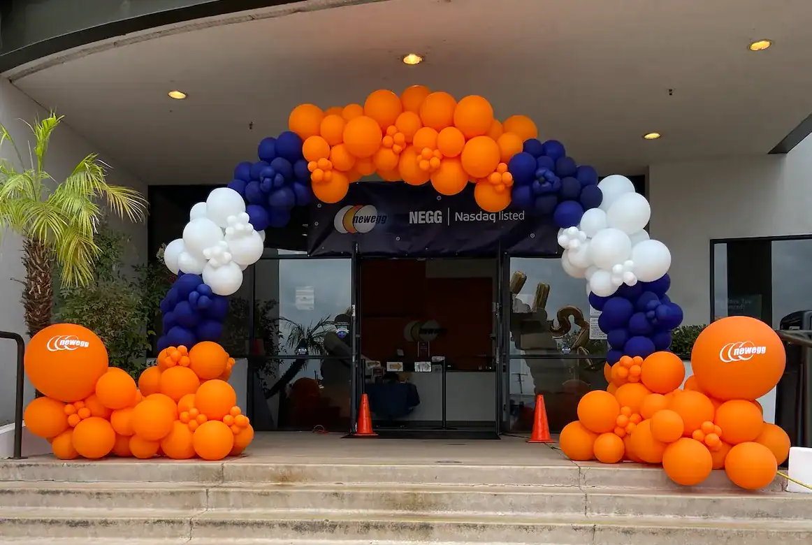 An archway made of orange, white, and blue balloons decorates the entrance of a building in Los Angeles, with orange Newegg-branded balloons on either side of the steps—showcasing impressive balloon installations.