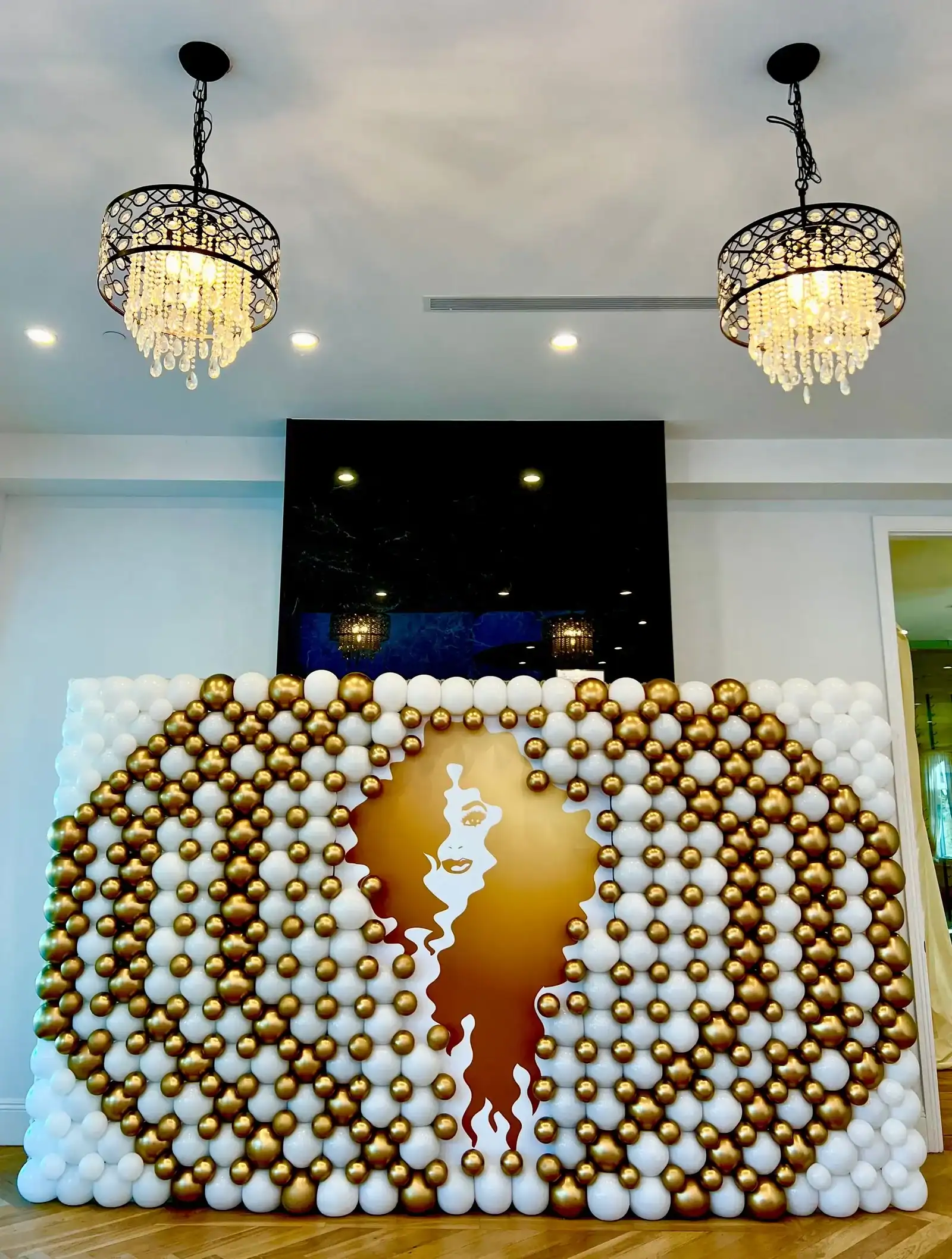 A decorative balloon wall with white and gold balloons, designed by a talented Los Angeles balloon artist, surrounds a gold silhouette of a woman's face and hair beneath two ornate chandeliers in a modern interior.