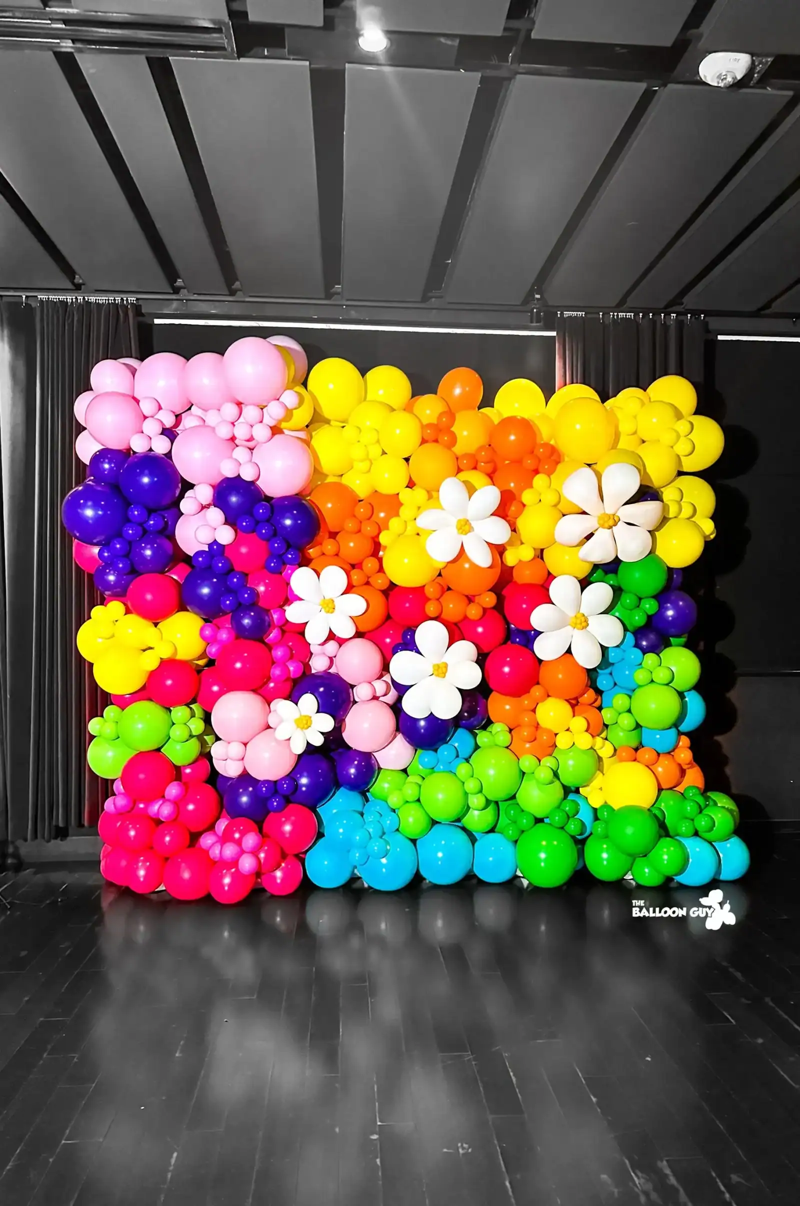 A colorful balloon wall with flowers made of balloons, crafted by a skilled balloon artist, stands in a rainbow pattern on a dark stage in Los Angeles.