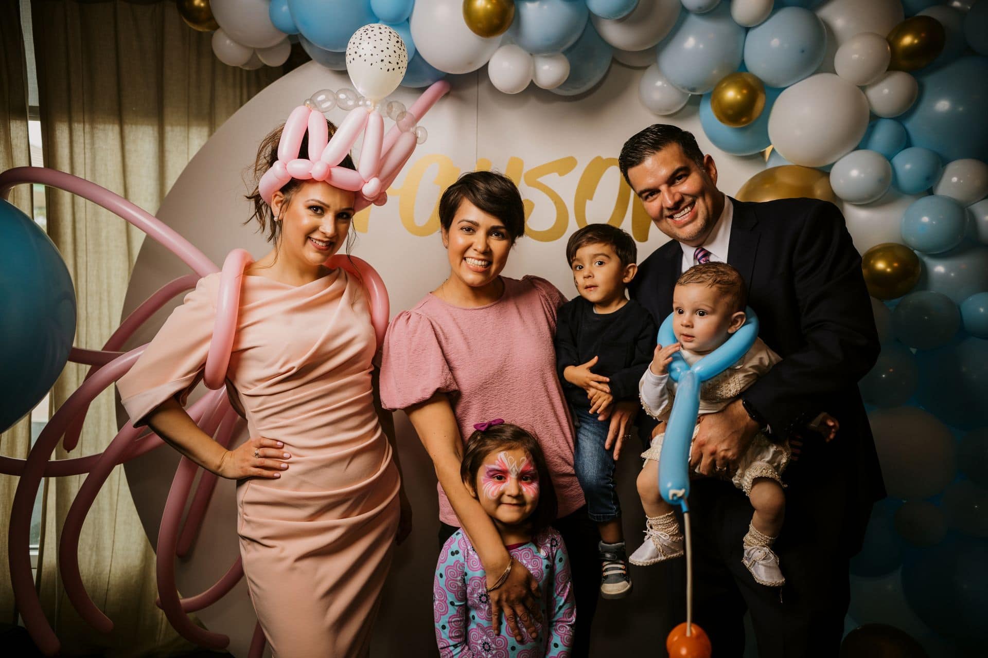 A group of five people, including a woman in a pink dress with balloon art, pose in front of a vibrant Los Angeles-inspired balloon installation. One child has face paint, while another holds a playful balloon sword.