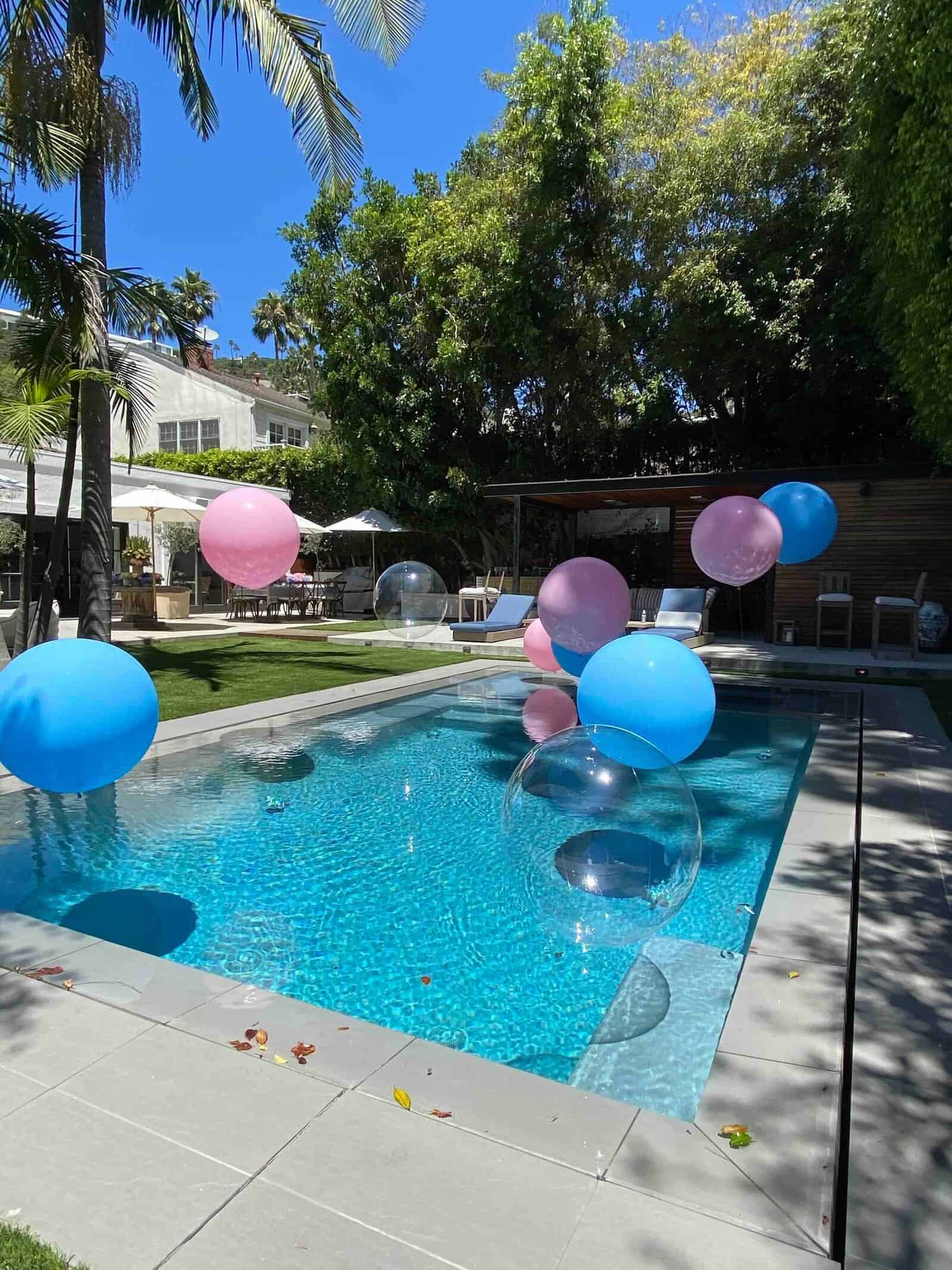 A swimming pool in Los Angeles surrounded by lounge chairs and trees, featuring vibrant balloon decor with large blue, pink, and clear balloons floating on the water.