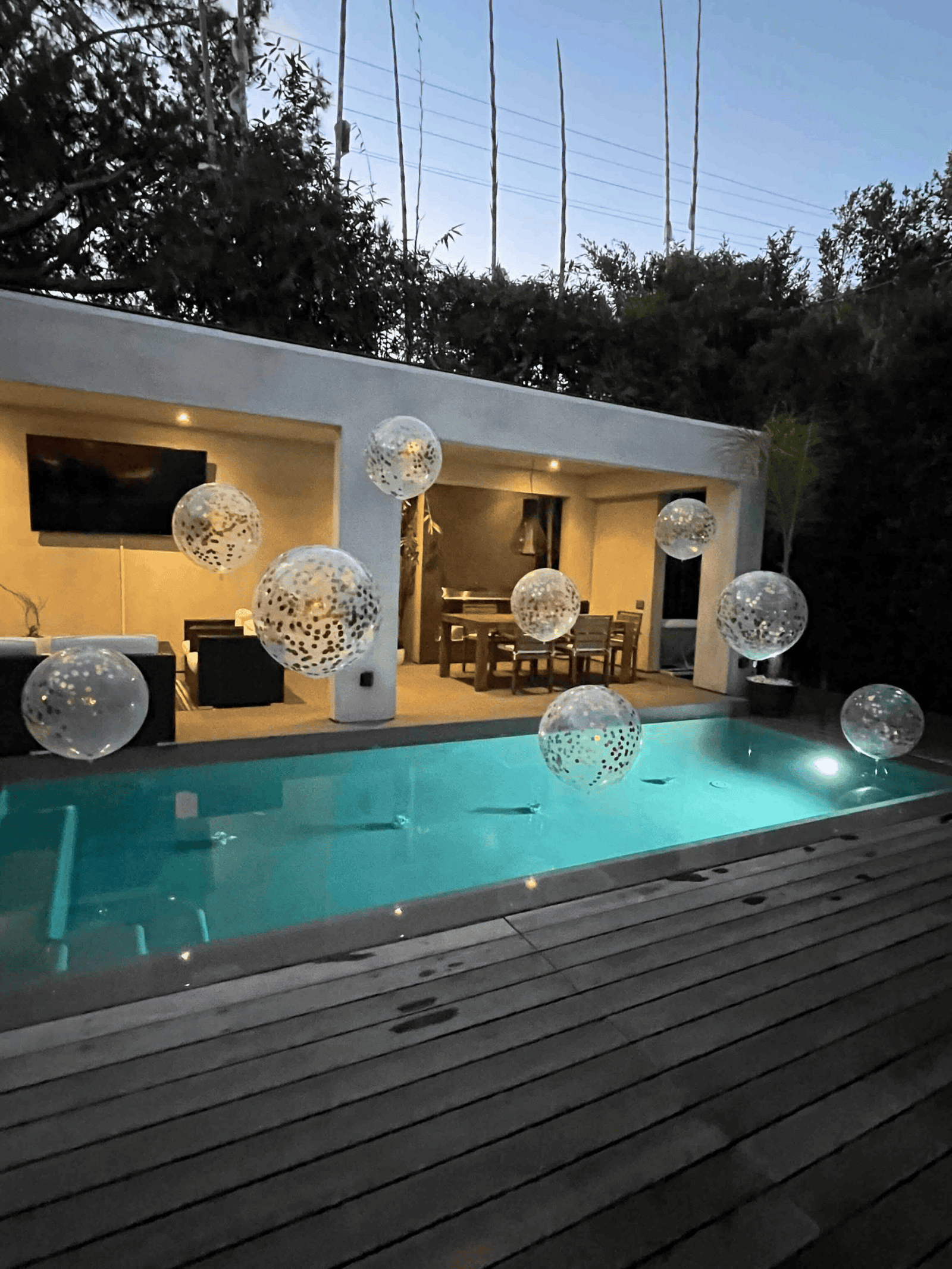 A modern backyard with a lit swimming pool, wooden deck, and a covered patio with a dining table. Elegant balloon decor from a talented balloon artist features transparent balloons floating over the pool in the evening light.