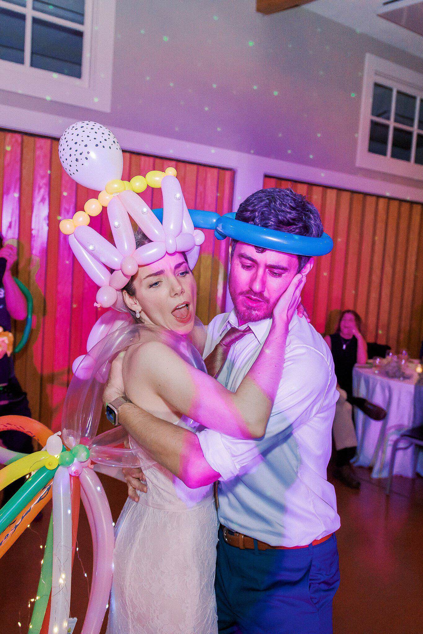 A bride and groom dance together at a wedding reception, wearing balloon crowns crafted by a balloon artist, with colorful balloon installations and guests seated in the background.