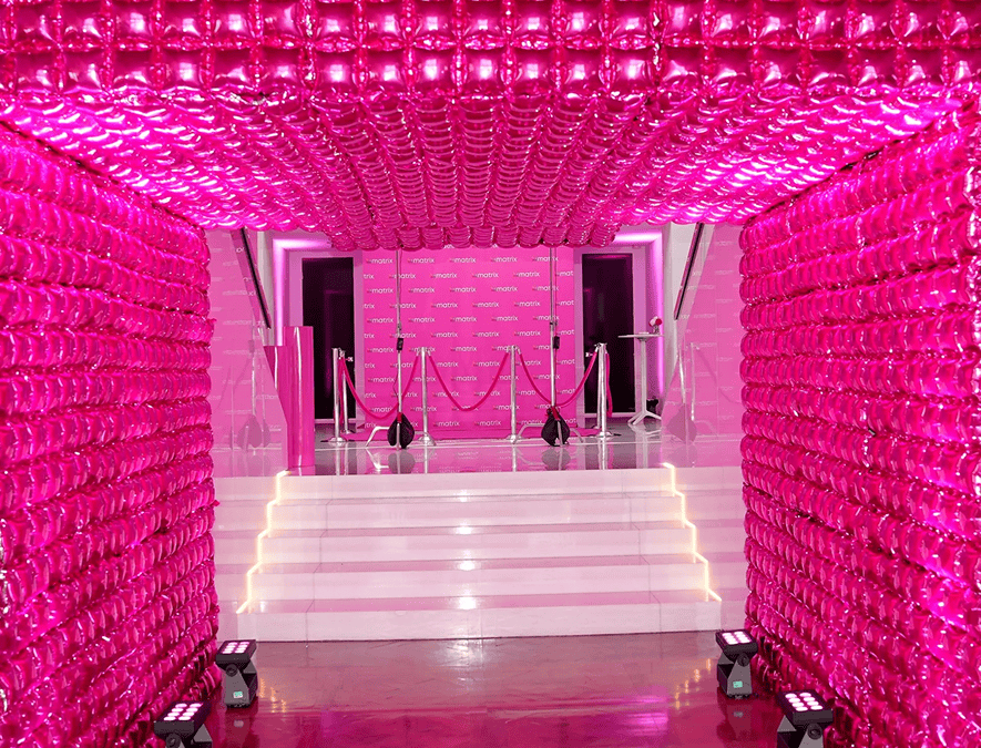 A hallway lined with shiny pink balloon decor leads to a staircase and a pink backdrop with ropes, surrounded by bright pink lighting.