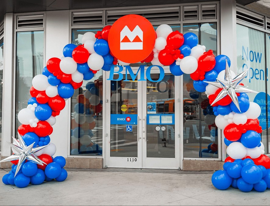A BMO bank entrance in Los Angeles decorated with a red, white, and blue balloon arch by a skilled balloon artist, silver star balloons, and "Now Open" signage visible inside.