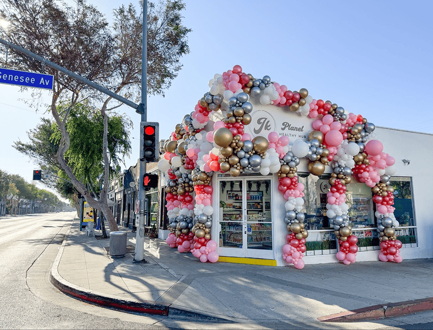 A white storefront on a street corner in Los Angeles is decorated with stunning balloon installations in pink, gold, white, and silver; a street sign reads "Genesee Av.