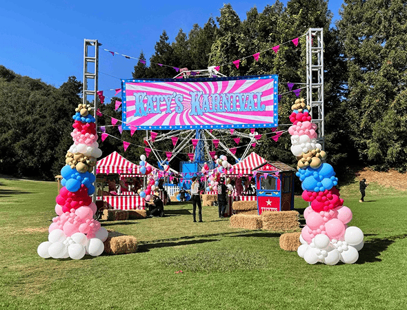 Outdoor carnival setup with a large "Katy's Karnival" sign, striped tents, eye-catching balloon installations, hay bales, and people enjoying the grassy field—perfect for a festive corporate event in Los Angeles with trees in the background.