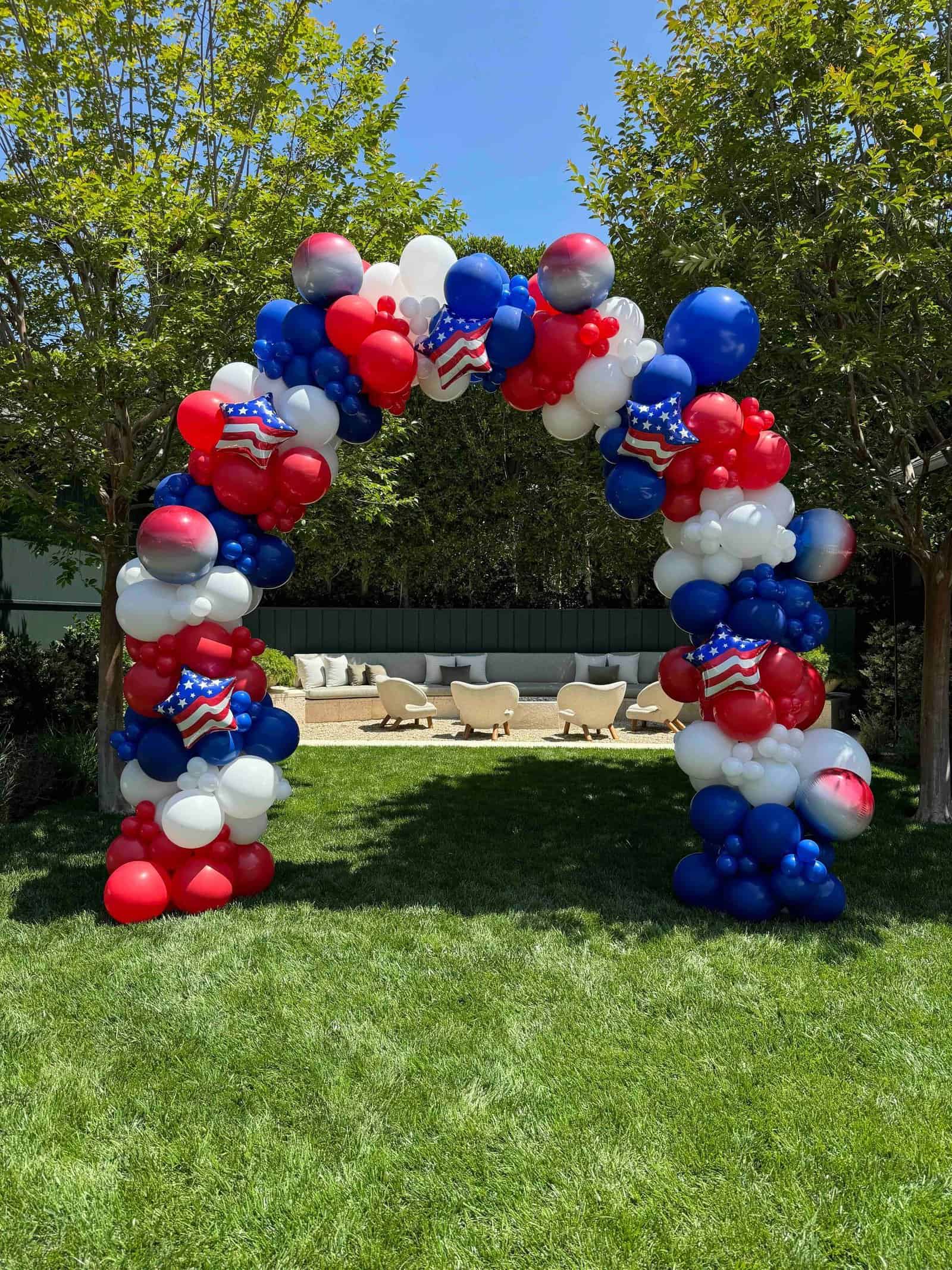 An arch made of red, white, and blue balloons with American flag patterns—crafted by a skilled balloon artist—stands on a grassy lawn in Los Angeles, with trees and outdoor seating visible in the background.