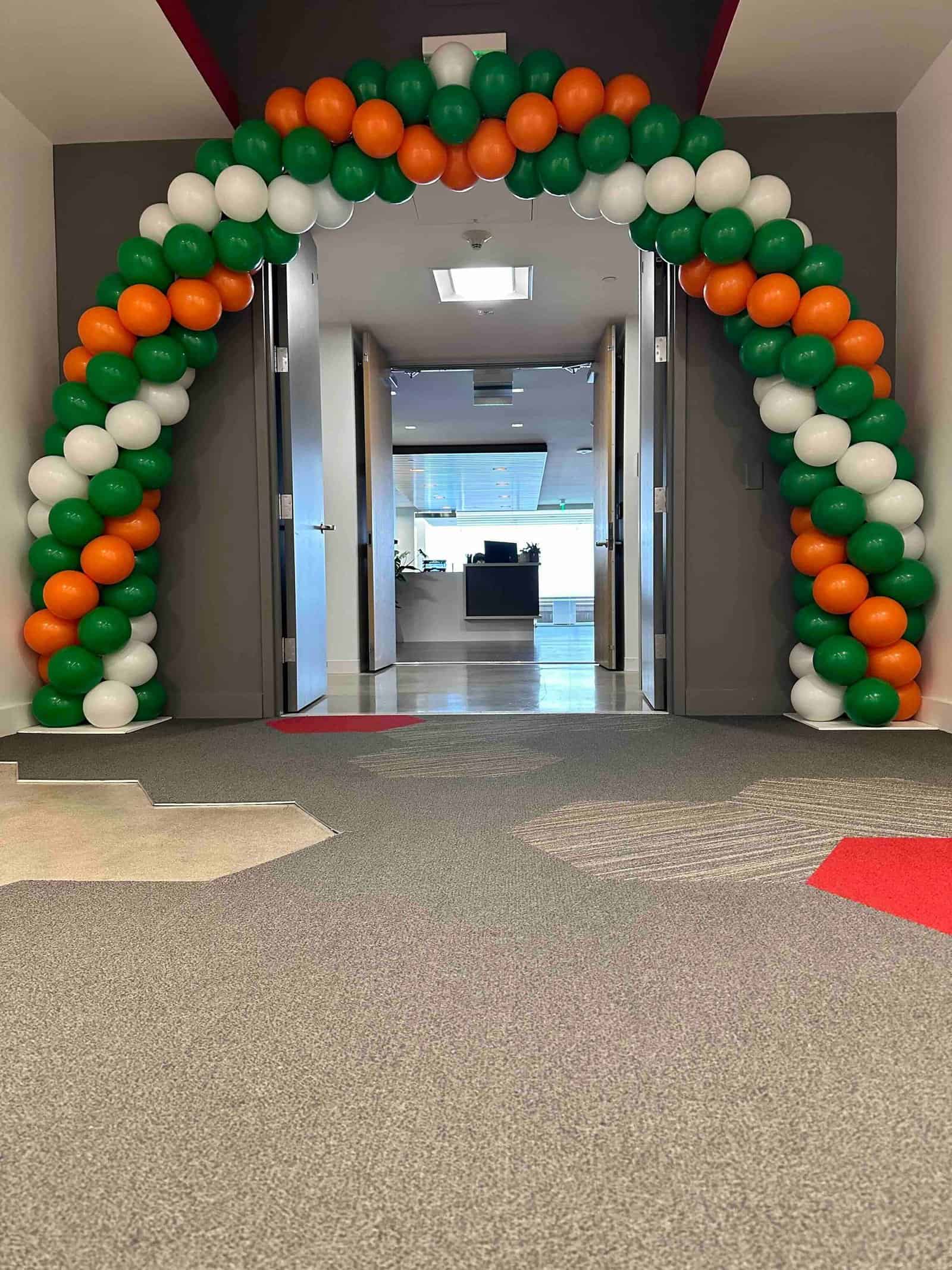A balloon artist crafted a stunning arch of green, orange, and white balloons to decorate the entrance to an office hallway for a corporate event.