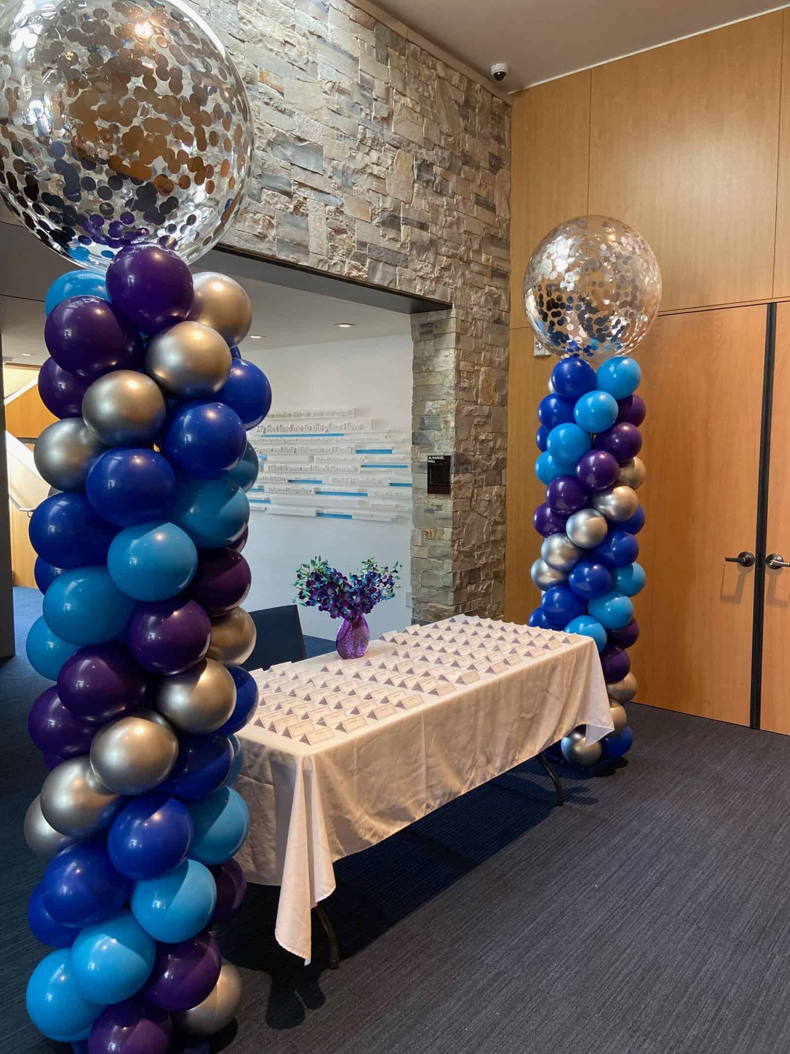 A table with a white cloth holds numerous place cards and a vase of purple flowers, flanked by stunning balloon decor columns crafted by a balloon artist, each topped with metallic confetti balloons.