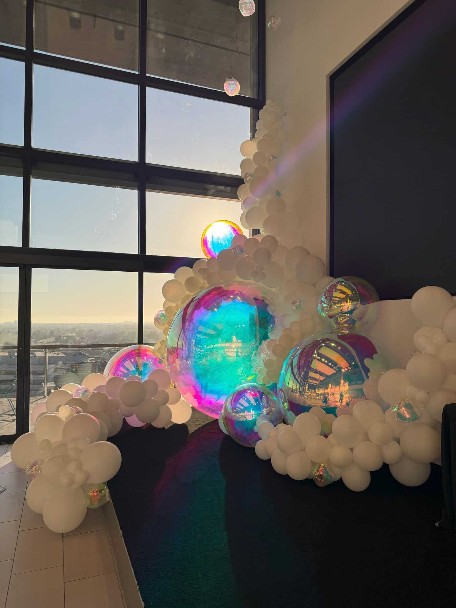 A decorative balloon installation featuring large iridescent and smaller white balloons is arranged by tall windows, with sunlight shining through—perfect for an elegant corporate event.