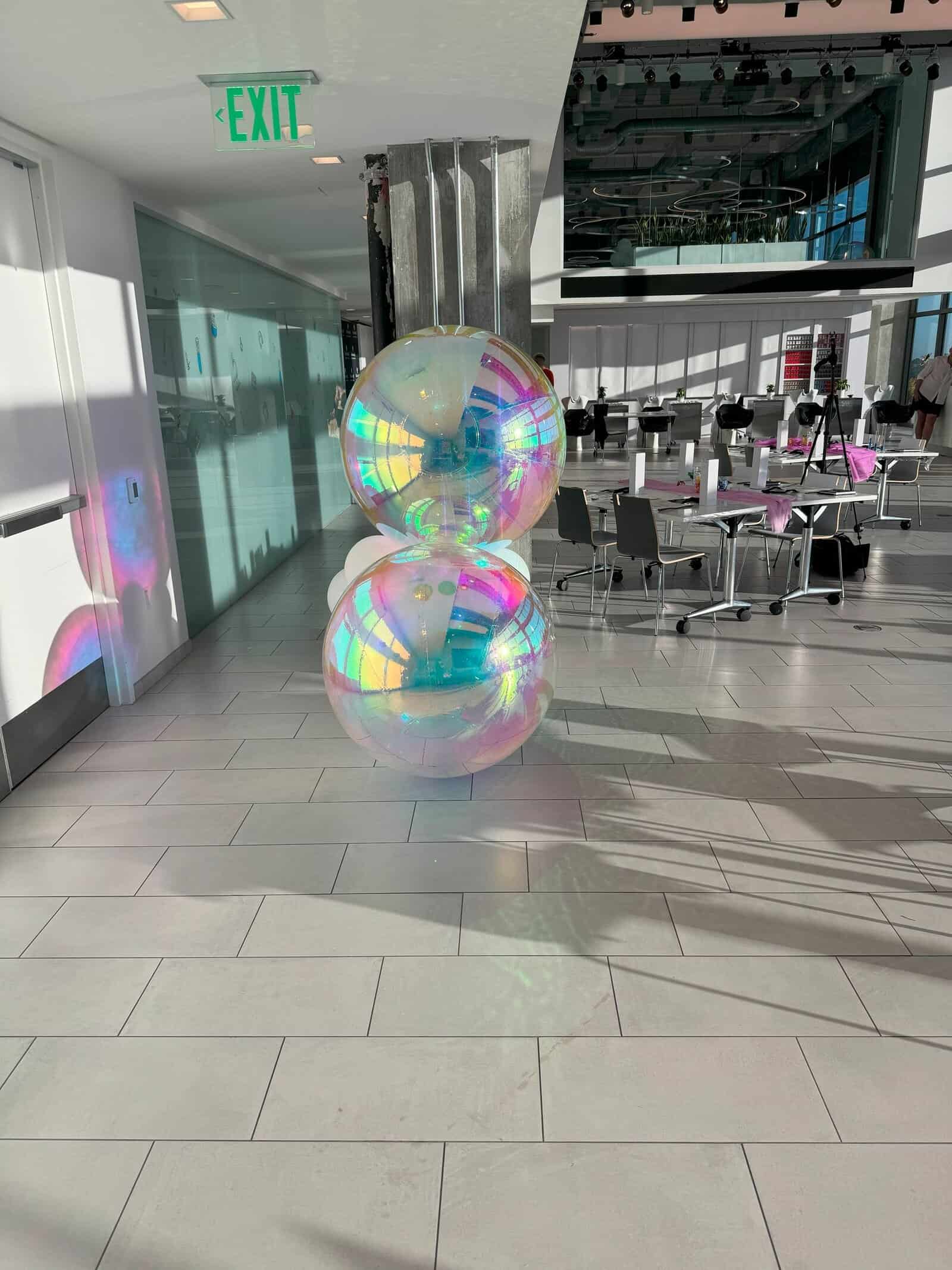 Two large iridescent balloons are stacked on top of each other as striking balloon decor in a sunlit, modern Los Angeles cafeteria with tables, chairs, and an EXIT sign overhead.