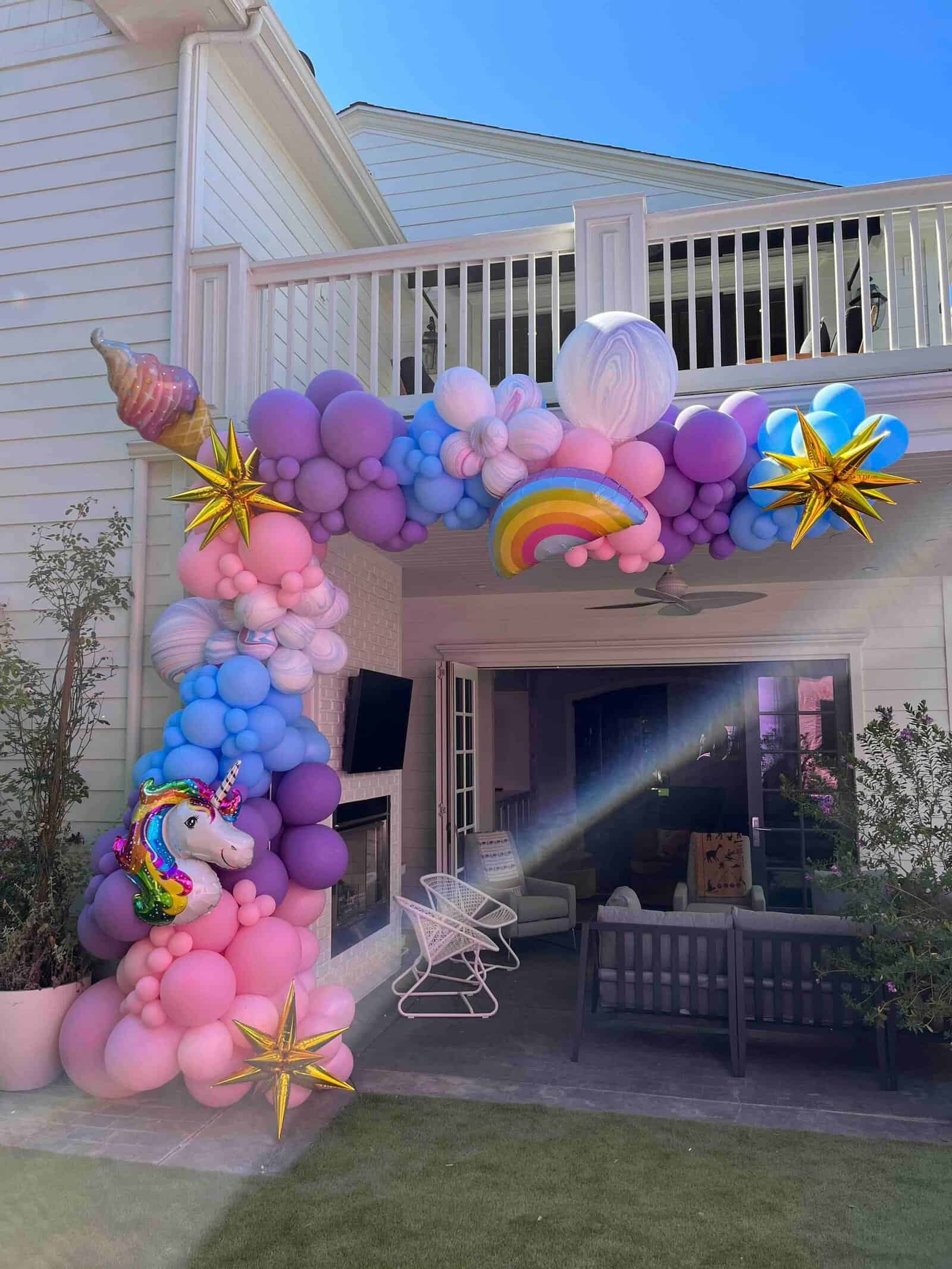 Colorful balloon arch with unicorn, rainbow, and star designs decorates the entrance of a patio at a white two-story house under a clear sky, showcasing creative balloon installations perfect for any festive or corporate event.