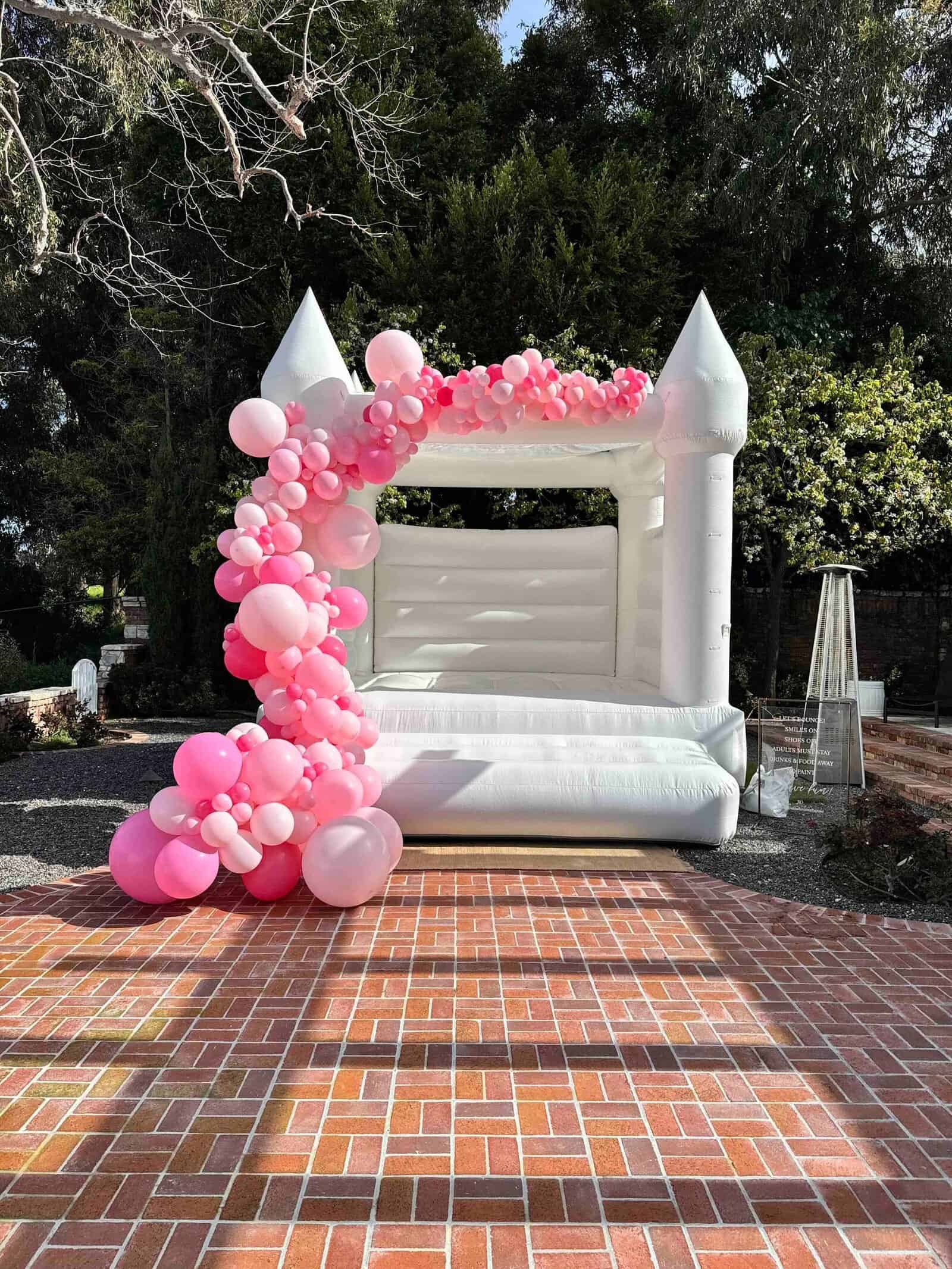 A white inflatable bounce house decorated with pink and white balloons stands on a brick patio outdoors in Los Angeles, surrounded by trees and greenery—perfect for any corporate event or celebration.
