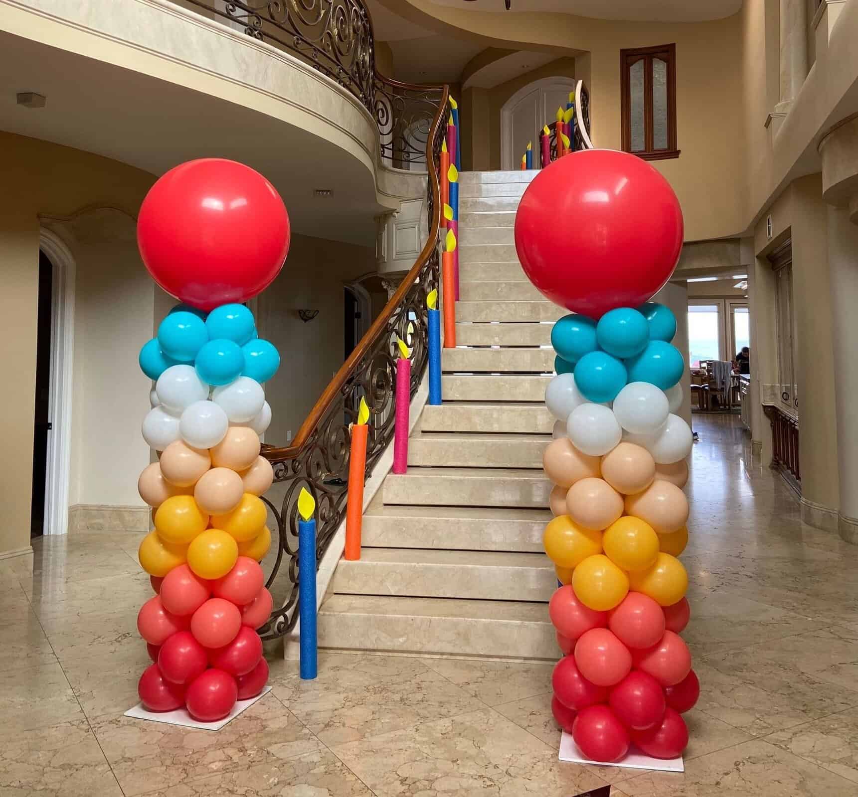 Two columns of colorful balloon installations with large red balloons on top flank a marble staircase in an indoor Los Angeles setting. The staircase is decorated with balloon arrangements resembling candles.