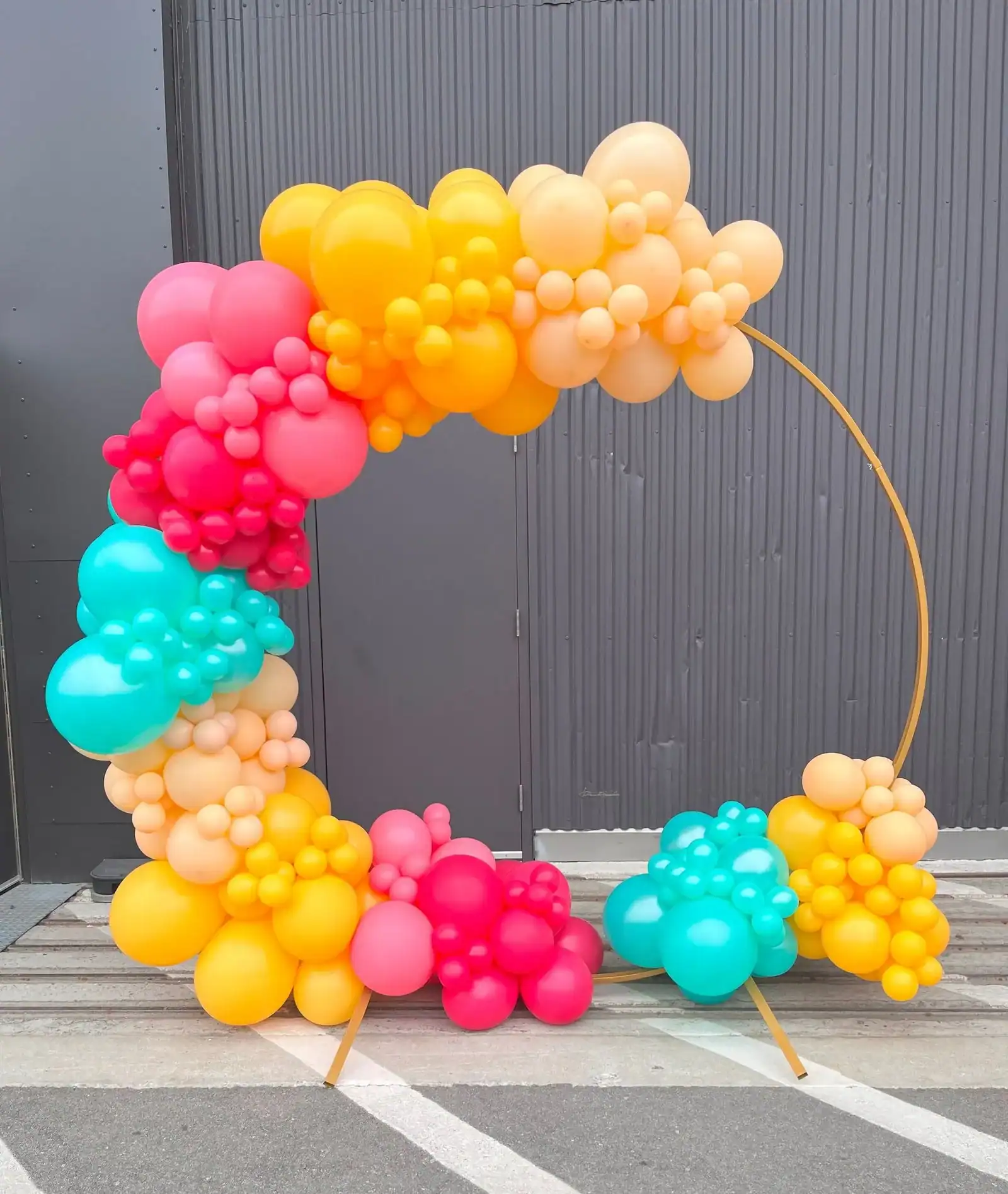 A large circular balloon arch with clusters of yellow, pink, orange, and teal balloons stands in front of a corrugated metal wall, perfect for adding vibrant flair to any Los Angeles corporate event.