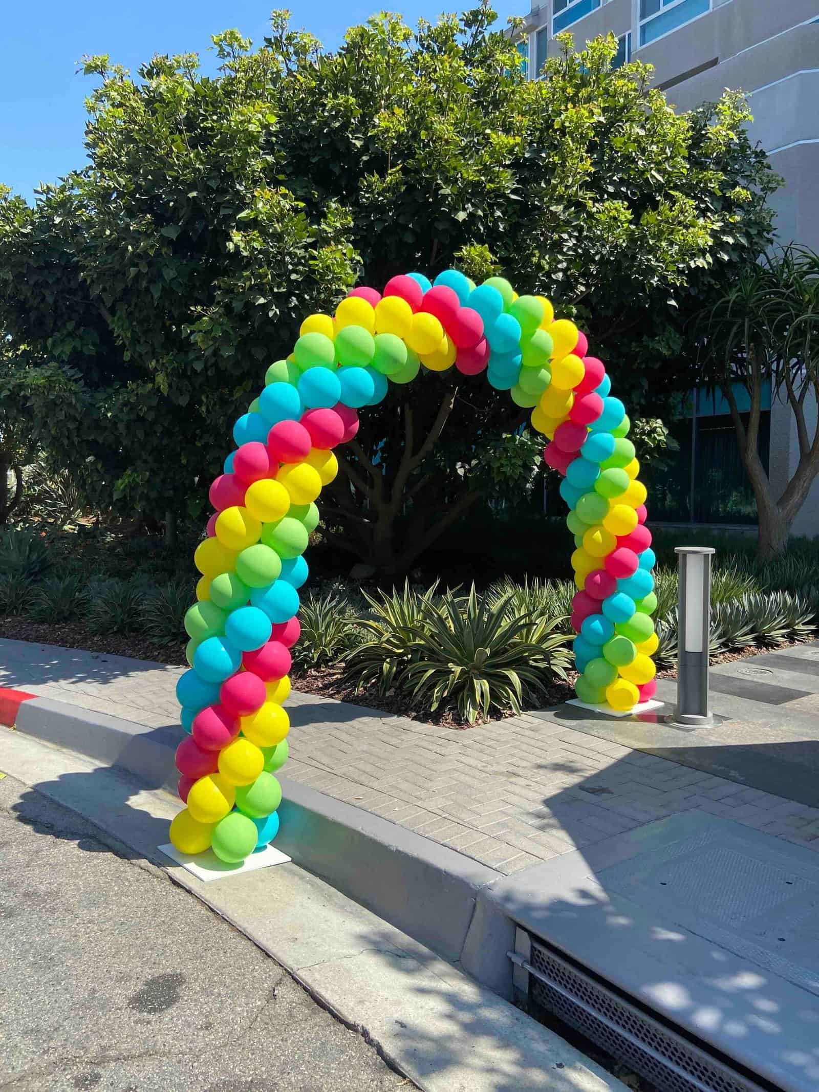 A colorful arch made of blue, yellow, red, and green balloons stands on a sidewalk near a landscaped area with trees and bushes, showcasing vibrant balloon decor.