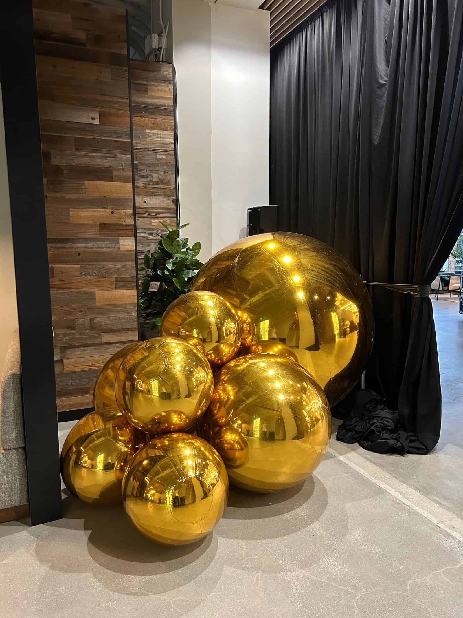 A cluster of large and small shiny gold balloons, arranged by a skilled balloon artist, sits on a concrete floor near a black curtain and wood-paneled wall in a modern indoor space.