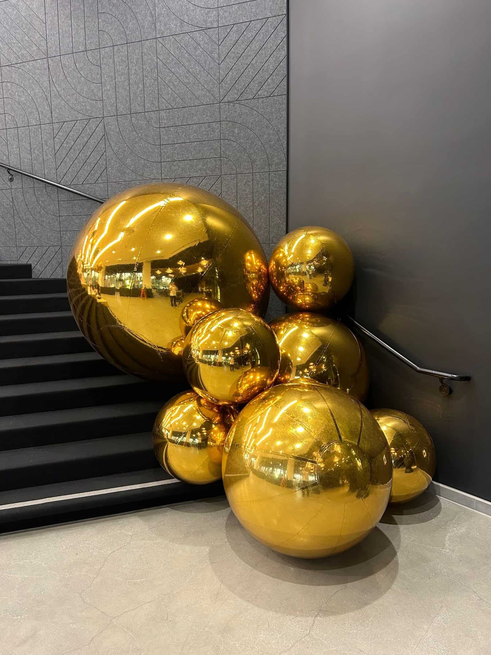 A cluster of large, shiny gold spheres creates striking balloon decor at the bottom of a staircase, next to a gray textured wall—perfect for any corporate event.