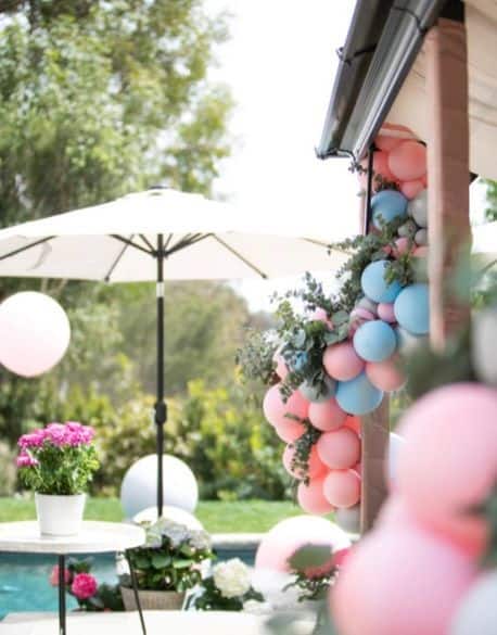 Outdoor scene in Los Angeles with pink and blue balloon installations decorating a structure, a white patio umbrella, and potted flowers on a table near the pool.